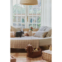 A cozy interior scene with a bed or daybed covered in beige and pink plaid bedding, styled with dog-shaped plush toys and pillows. A window with white frames and sheer curtains is behind the bed, and a basket with a dog toy sits on the floor nearby.