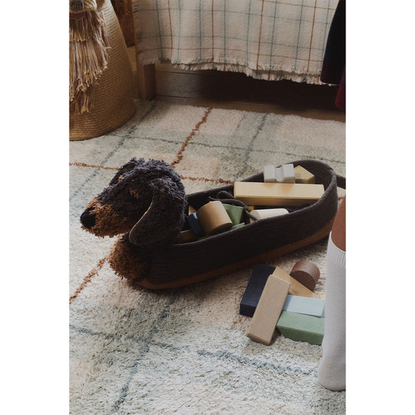 Dog basket  resembling a dachshund filled with toys on a checkered floor.