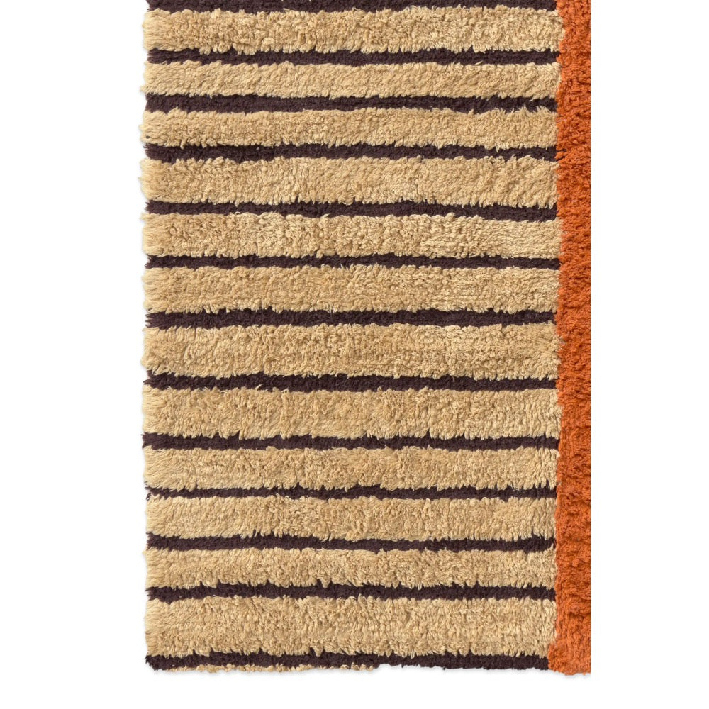 A close-up of a textured shag rug featuring horizontal beige and dark brown stripes, with a solid rust-orange border on the right side.