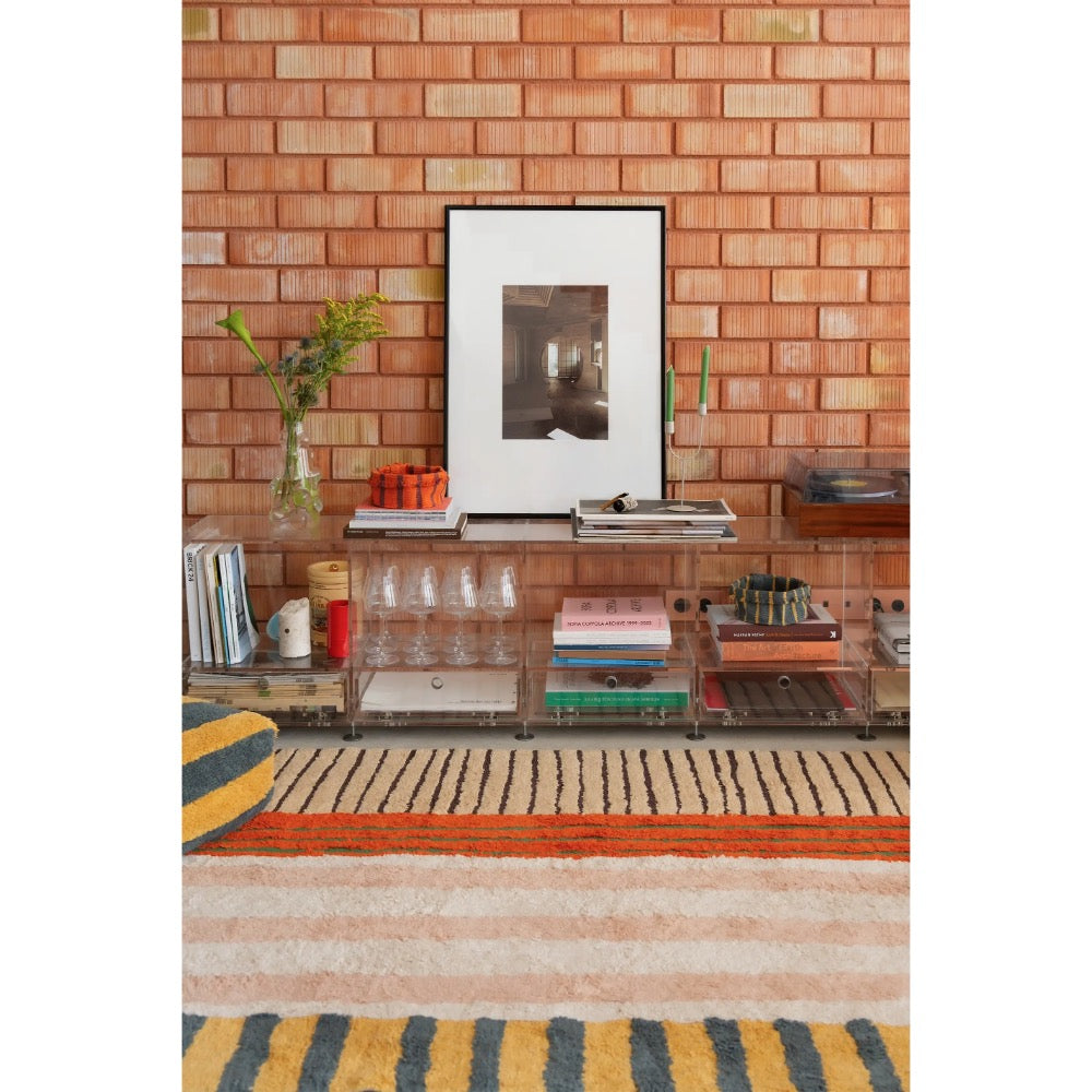 A clear acrylic bar cart with multiple tiers and open shelving, styled with books, glasses, and a vase in a living room setting with a brick wall background.