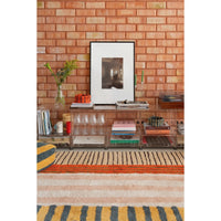 A clear acrylic bar cart with multiple tiers and open shelving, styled with books, glasses, and a vase in a living room setting with a brick wall background.