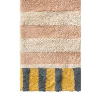A close-up of a plush shag rug with horizontal stripes in dusty pink, cream, and mustard yellow with dark gray vertical stripes at the bottom.
