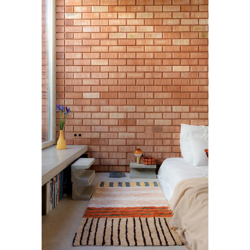 A styled bedroom interior featuring a prominent warm terracotta brick wall, a bed with white bedding, a built-in bench with books and a yellow vase, and two patterned rugs on the floor.