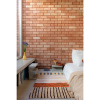 A styled bedroom interior featuring a prominent warm terracotta brick wall, a bed with white bedding, a built-in bench with books and a yellow vase, and two patterned rugs on the floor.