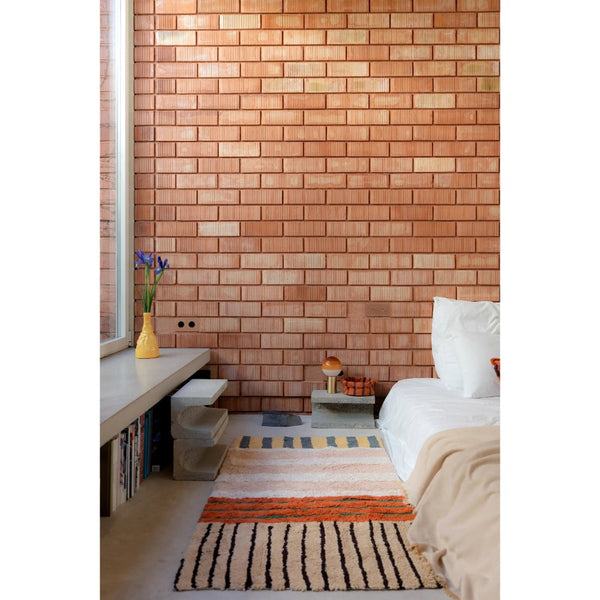 A styled bedroom interior featuring a prominent warm terracotta brick wall, a bed with white bedding, a built-in bench with books and a yellow vase, and two patterned rugs on the floor.