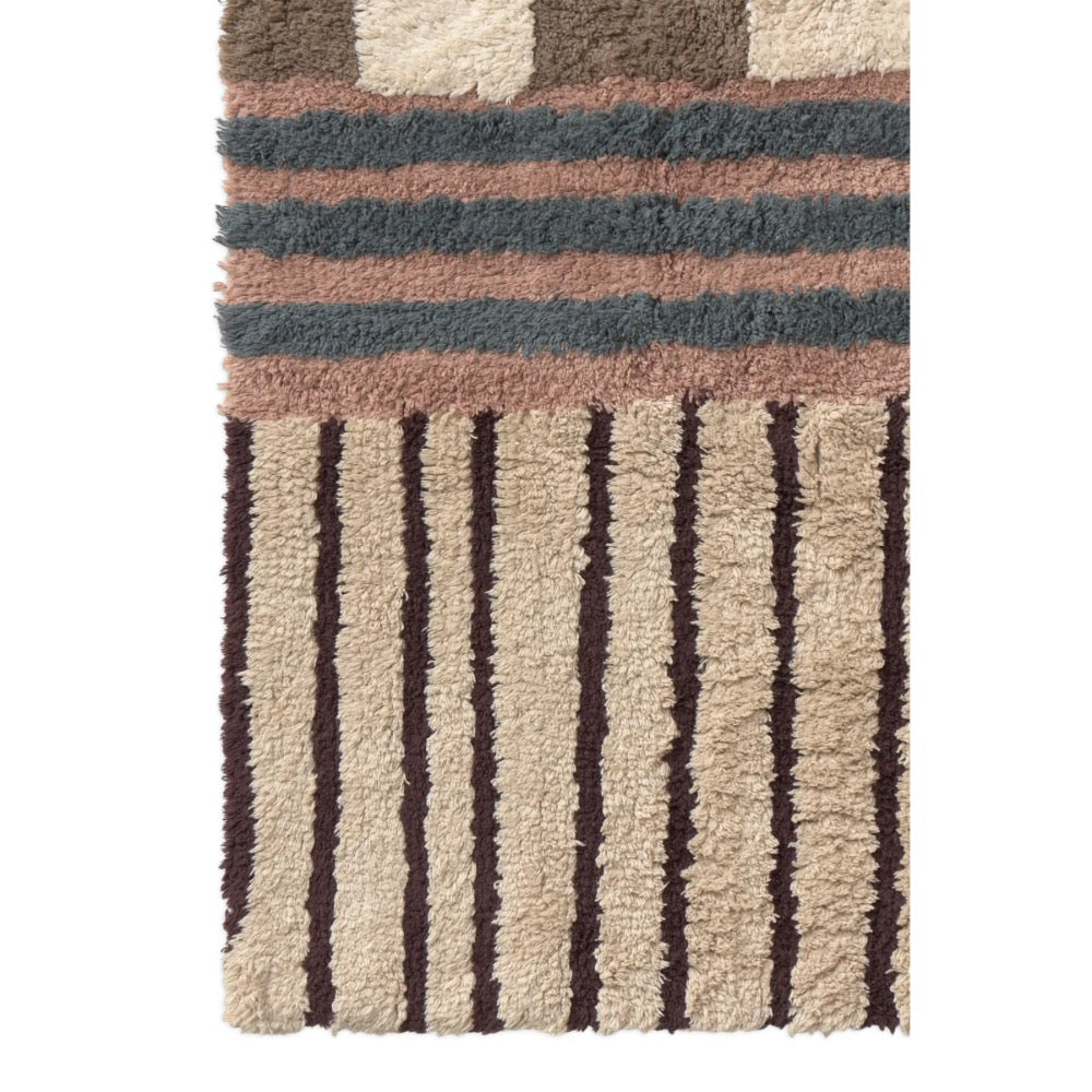 A close-up of a textured shag rug with a geometric pattern, featuring horizontal stripes in dusty pink, slate blue, and brown above vertical dark brown stripes on a beige background.