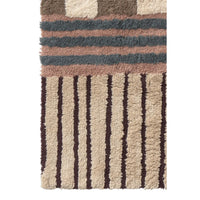 A close-up of a textured shag rug with a geometric pattern, featuring horizontal stripes in dusty pink, slate blue, and brown above vertical dark brown stripes on a beige background.
