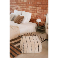 A cream bouclé pouf ottoman with black vertical stripes sits in a modern bedroom setting in front of a bed with white linens and a brick-patterned accent wall.