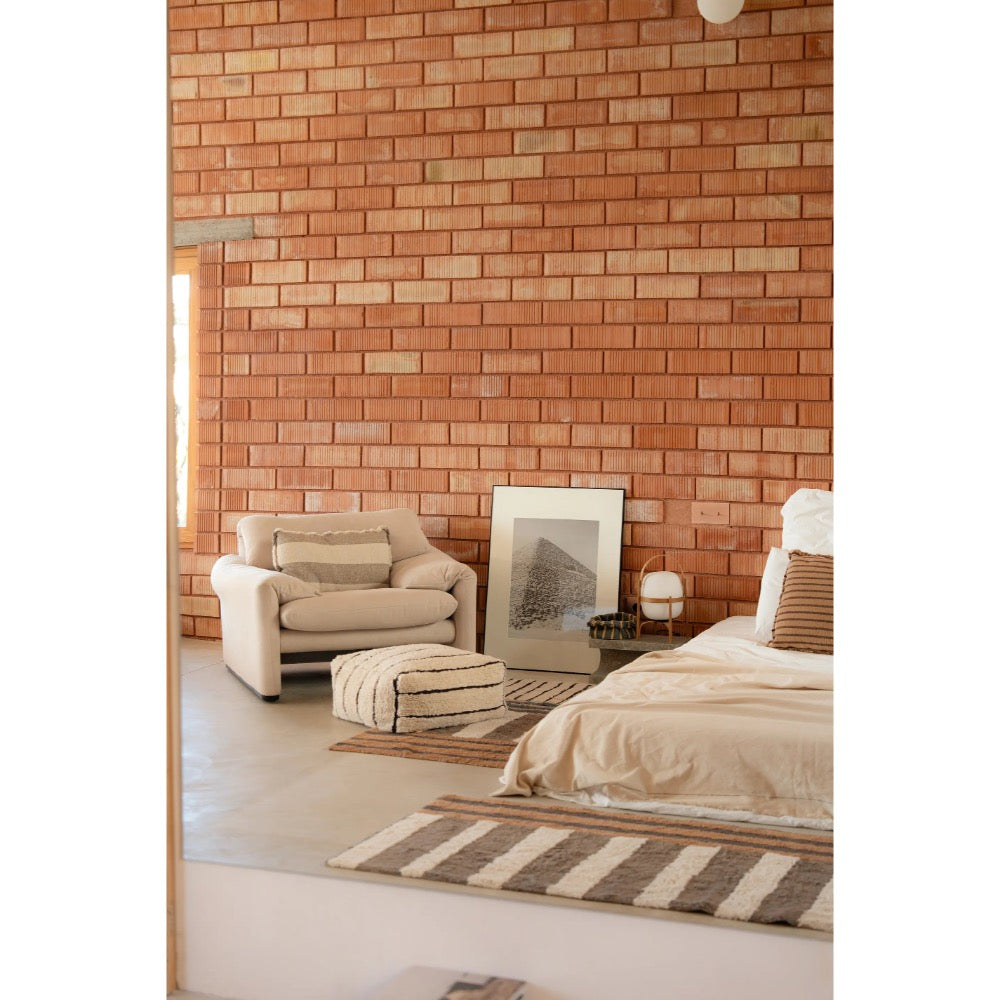 A styled bedroom interior featuring a terracotta brick accent wall, a cream armchair, a striped rug, a striped pouf, and a bed with neutral linens.