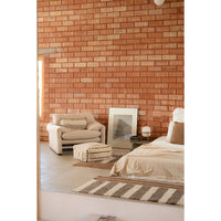 A styled bedroom interior featuring a terracotta brick accent wall, a cream armchair, a striped rug, a striped pouf, and a bed with neutral linens.