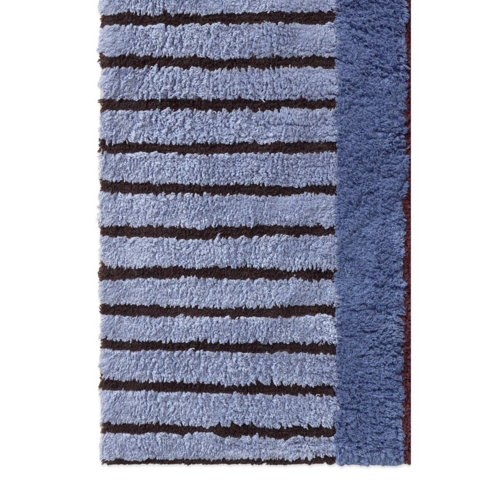 A close-up of a textured shag rug featuring horizontal stripes in light blue and dark brown, with a solid medium blue border on the right side.