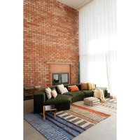 A modern living room featuring a dark green sectional sofa with patterned throw pillows, set on a striped blue, beige, and brown area rug in front of a brick wall and large white window curtains.