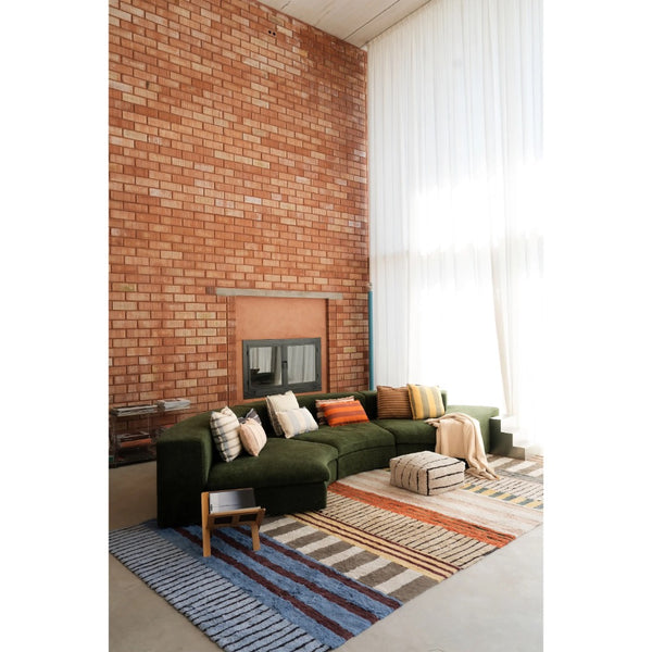 A modern living room featuring a dark green sectional sofa with patterned throw pillows, set on a striped blue, beige, and brown area rug in front of a brick wall and large white window curtains.