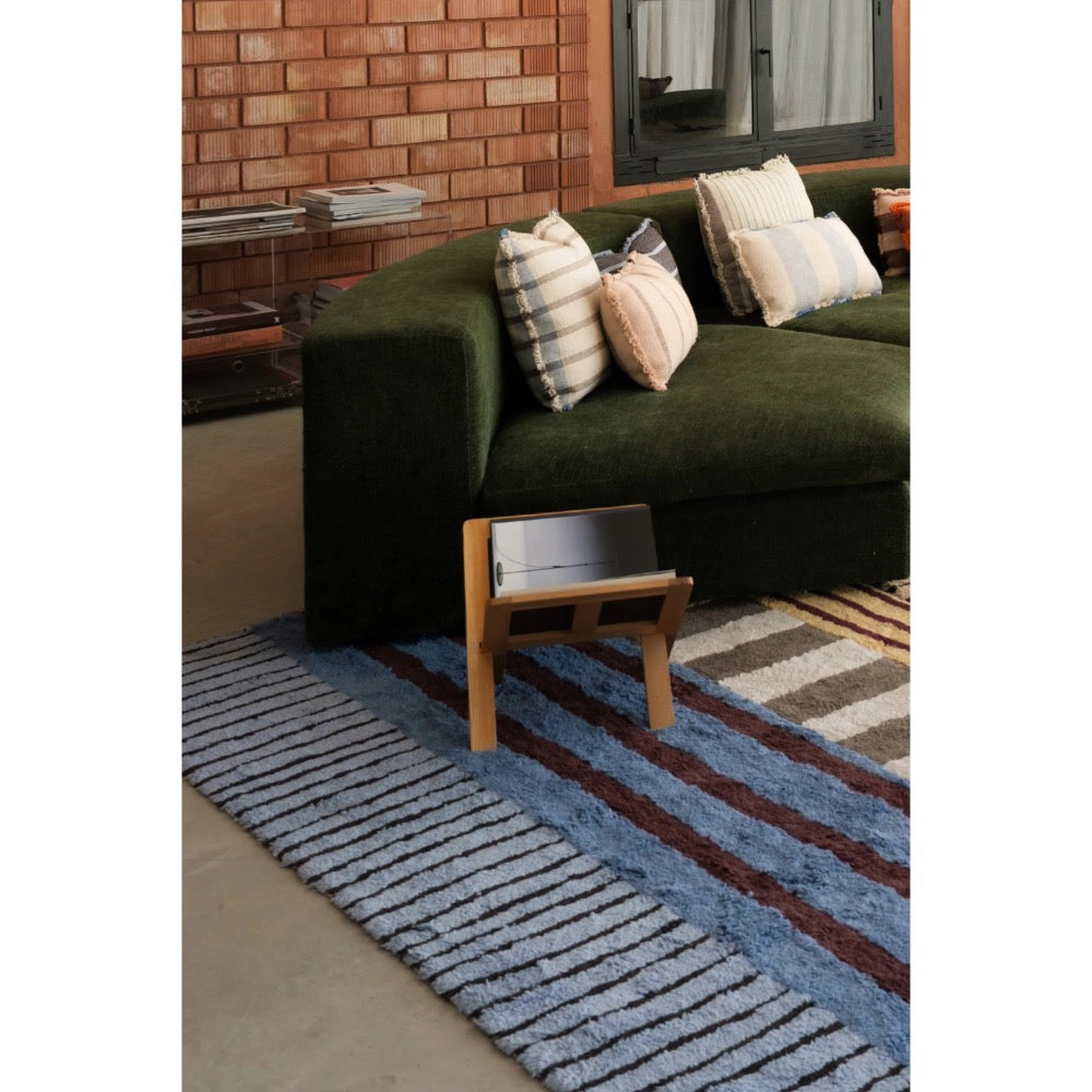 A blue and brown striped shag area rug styled in a modern living room setting with a dark green sofa, striped throw pillows, and a small wooden magazine stand.