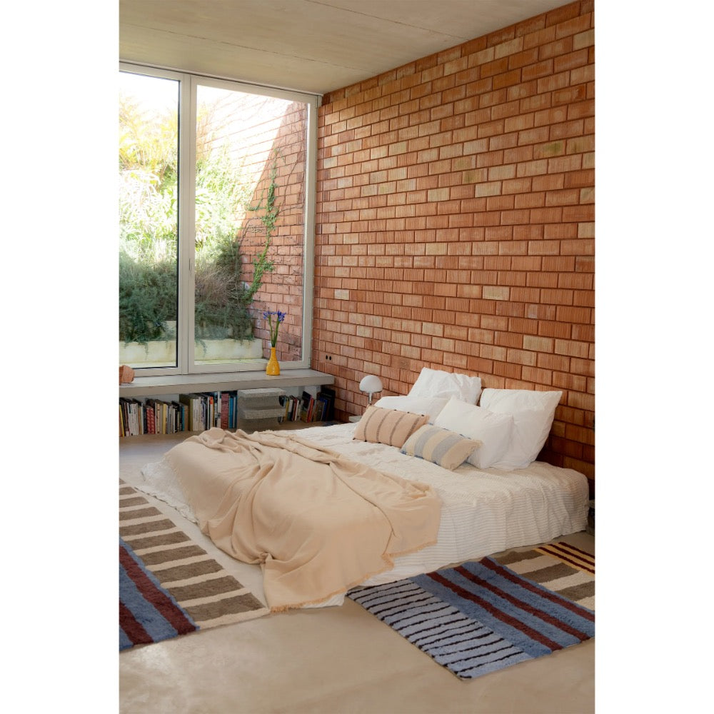 A low bed with white bedding and a beige throw blanket in a modern bedroom featuring a brick accent wall, a large window, and striped rugs.