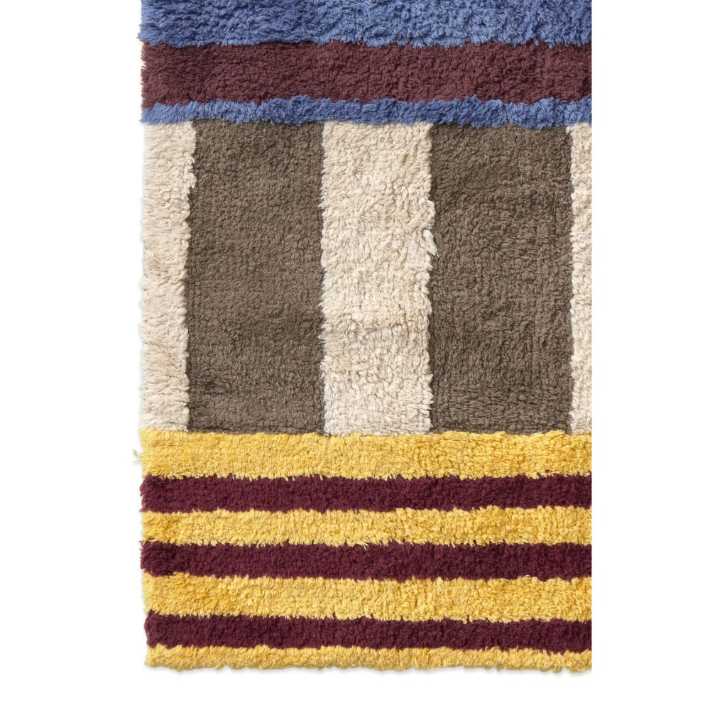 A close-up of a textured shag rug with a geometric pattern featuring vertical stripes in brown, cream, and gray, bordered by blue and burgundy at the top, and yellow and burgundy stripes at the bottom.
