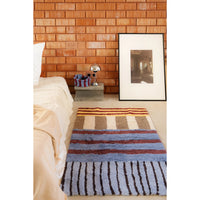 A striped shag area rug in blue, brown, beige, and white lies on a light-colored floor in a bedroom setting, with a bed, side table, lamp, and framed picture against a wood shingle wall.