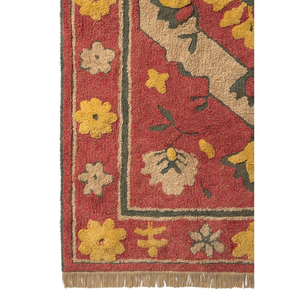 Close-up of a patterned area rug showing a red field with beige, yellow, and dark green floral motifs and fringe along the bottom edge.