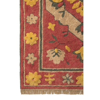 Close-up of a patterned area rug showing a red field with beige, yellow, and dark green floral motifs and fringe along the bottom edge.
