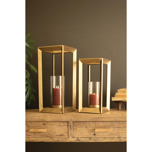 Two gold-toned metal lantern candle holders with clear glass sides and red pillar candles, displayed on a rustic wooden table against a dark brown wall.