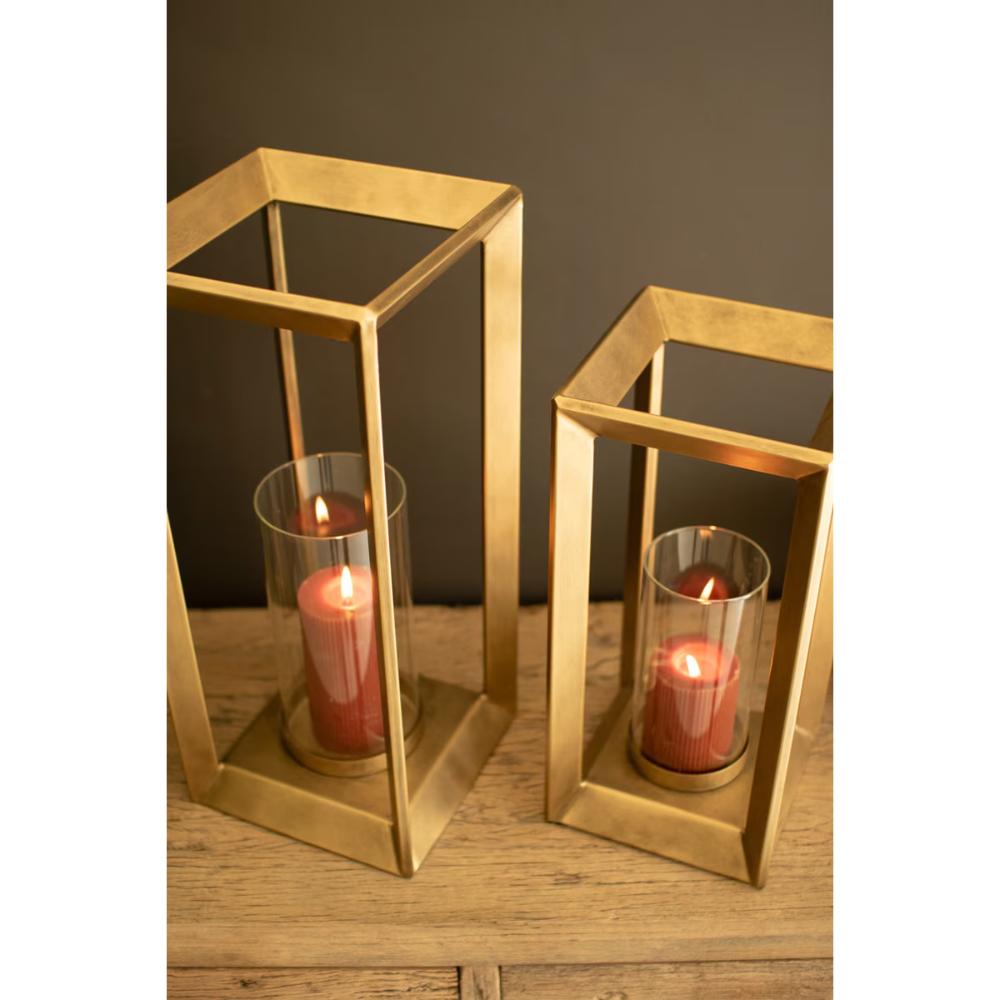 Two gold-toned open cube lantern candle holders with clear glass sides and lit red pillar candles, displayed on a wooden surface against a dark gray background.