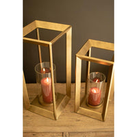 Two gold-toned open cube lantern candle holders with clear glass sides and lit red pillar candles, displayed on a wooden surface against a dark gray background.