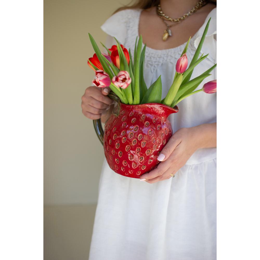 Person holding a red strawberry-shaped vase filled with flowers against a plain background