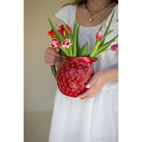 Person holding a red strawberry-shaped vase filled with flowers against a plain background
