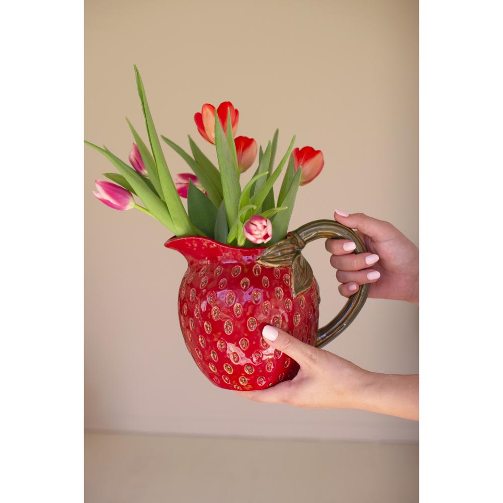 Red pitcher filled with flowers held by a hand against a beige background