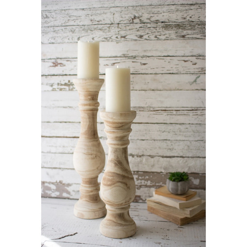 Two Kalalou Hand Carved Wooden Candle Stands with white candles complement books and a small plant on a rustic surface.