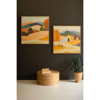 Two abstract landscape paintings on a dark wall with a woven ottoman and plant below.