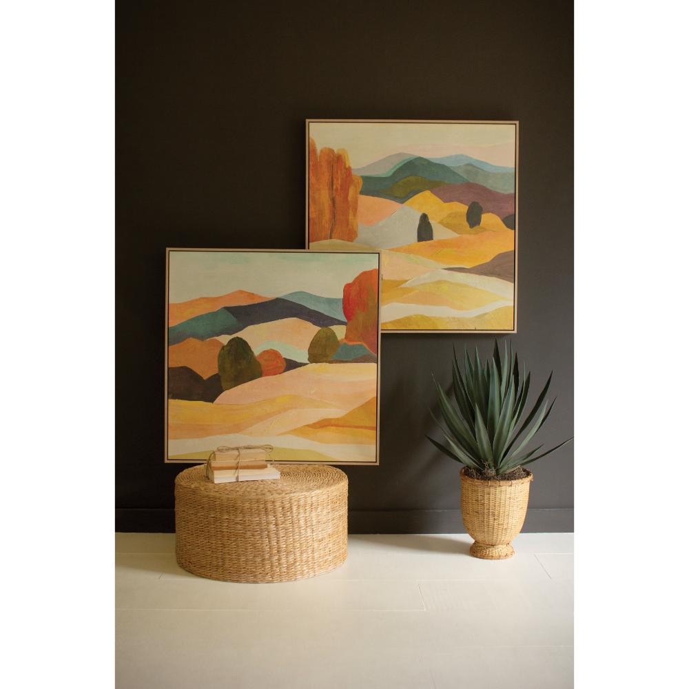 Two landscape paintings on a dark wall with a woven ottoman and plant in the foreground.