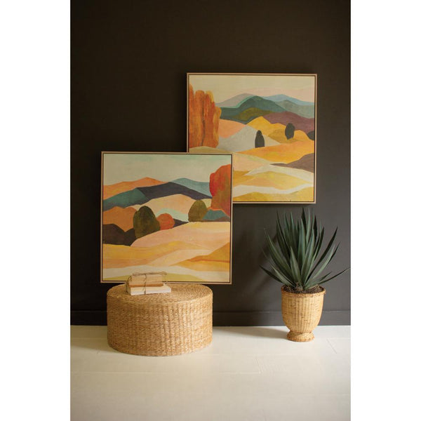 Two landscape paintings on a dark wall with a woven ottoman and plant in the foreground.
