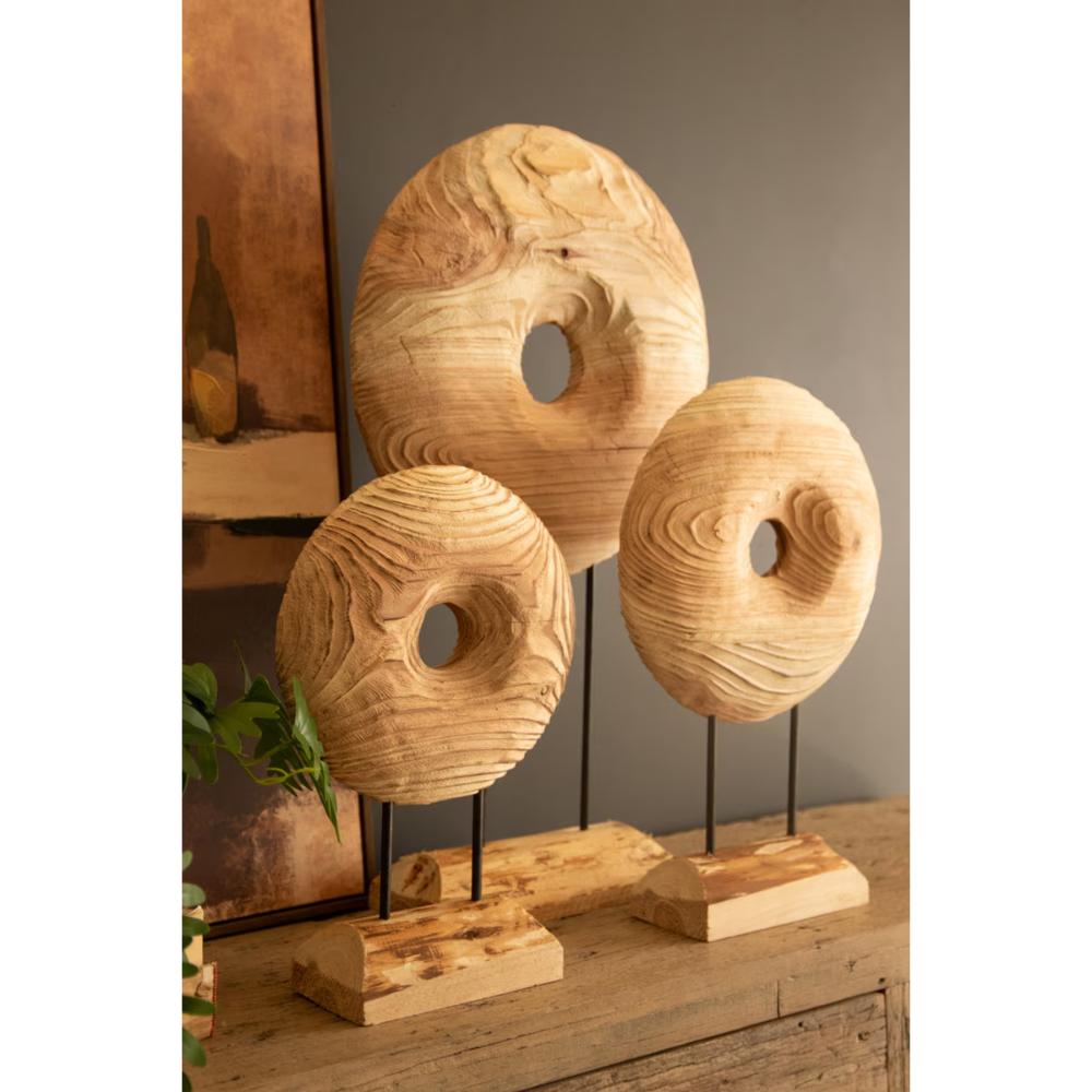 Three carved wooden donut sculptures with natural swirling grain, displayed on black metal stands with rectangular wooden bases on a rustic wooden table.