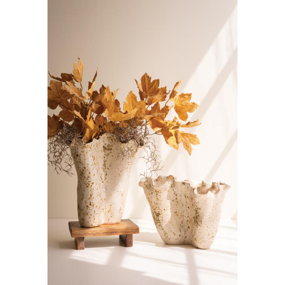 Two textured ceramic vases with a scalloped rim, one tall on a small wooden stand and one short, both filled with dried golden-brown leaves, set against a warm beige wall with soft window shadows.