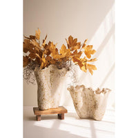 Two textured ceramic vases with a scalloped rim, one tall on a small wooden stand and one short, both filled with dried golden-brown leaves, set against a warm beige wall with soft window shadows.
