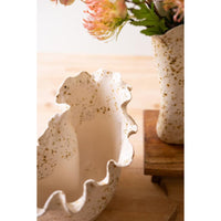 A close-up of a speckled ceramic bowl with a wavy rim, shown on a wooden surface next to another matching vase holding flowers.