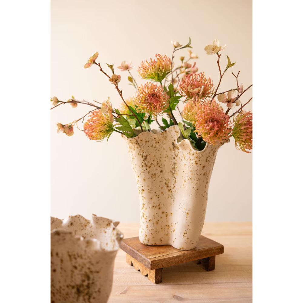 A ceramic double-vase flower holder with a speckled cream finish, filled with dried orange and pink flowers, displayed on a small wooden riser against a warm beige background.
