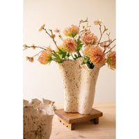 A ceramic double-vase flower holder with a speckled cream finish, filled with dried orange and pink flowers, displayed on a small wooden riser against a warm beige background.