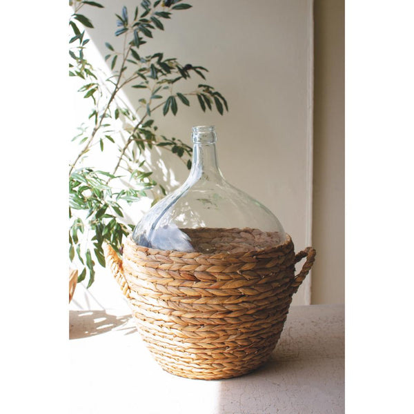 Clear glass demijohn in a woven basket with a plant in the background