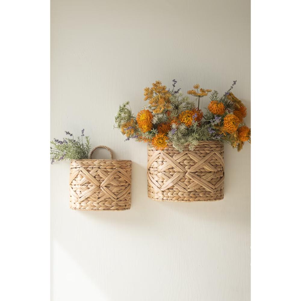 Two woven natural-fiber wall baskets hanging on a light neutral wall, with dried orange and yellow flowers arranged in them.