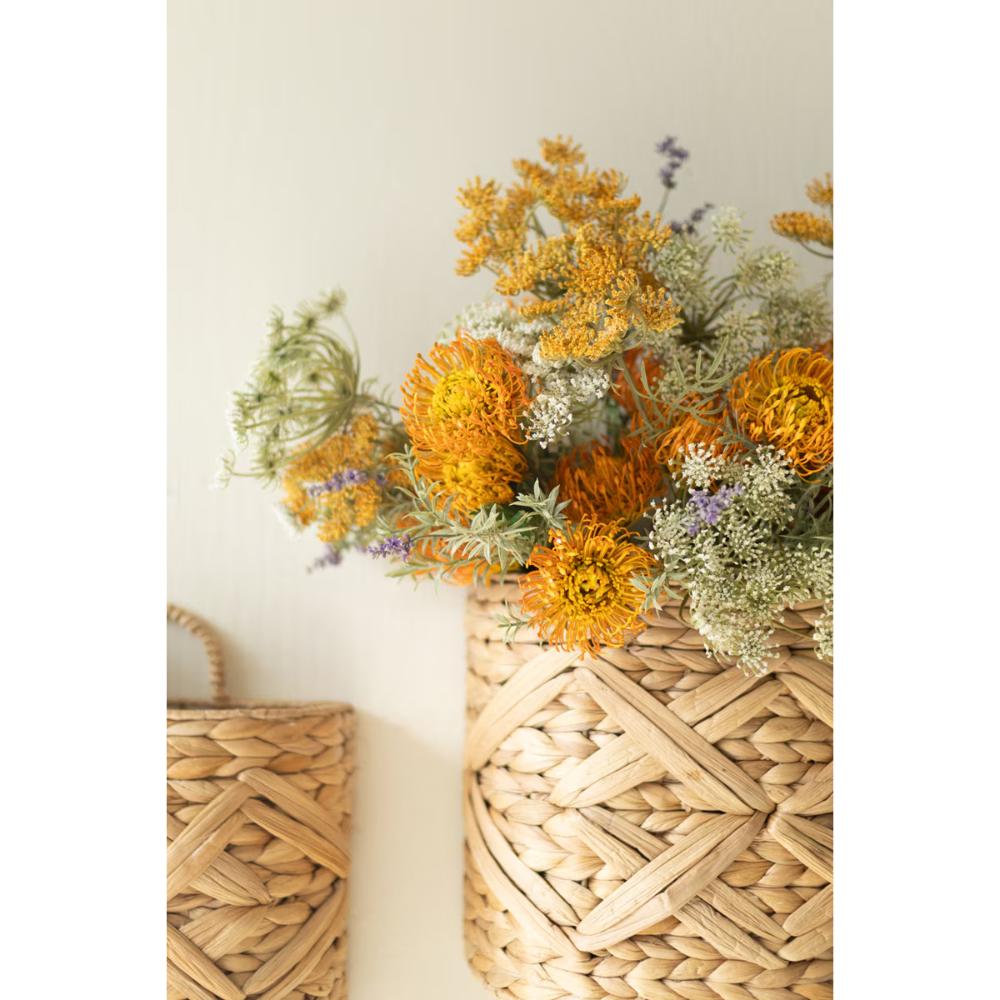A woven natural-fiber basket holding a bouquet of dried orange, yellow, and cream flowers against a plain light beige wall.