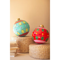 Two colorful Christmas ornament balls on a beige background. One ball is red with pink bands and green and yellow polka dots sitting on a round rattan riser, and the other is turquoise with yellow and purple polka dots and gold trim sitting on a round rattan riser