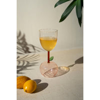 An Apple Base Wine Glass from Kalalou, with orange liquid and a leaf, sits on a table next to two lemons casting leaf shadows.