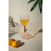 An Apple Base Wine Glass from Kalalou, with orange liquid and a leaf, sits on a table next to two lemons casting leaf shadows.