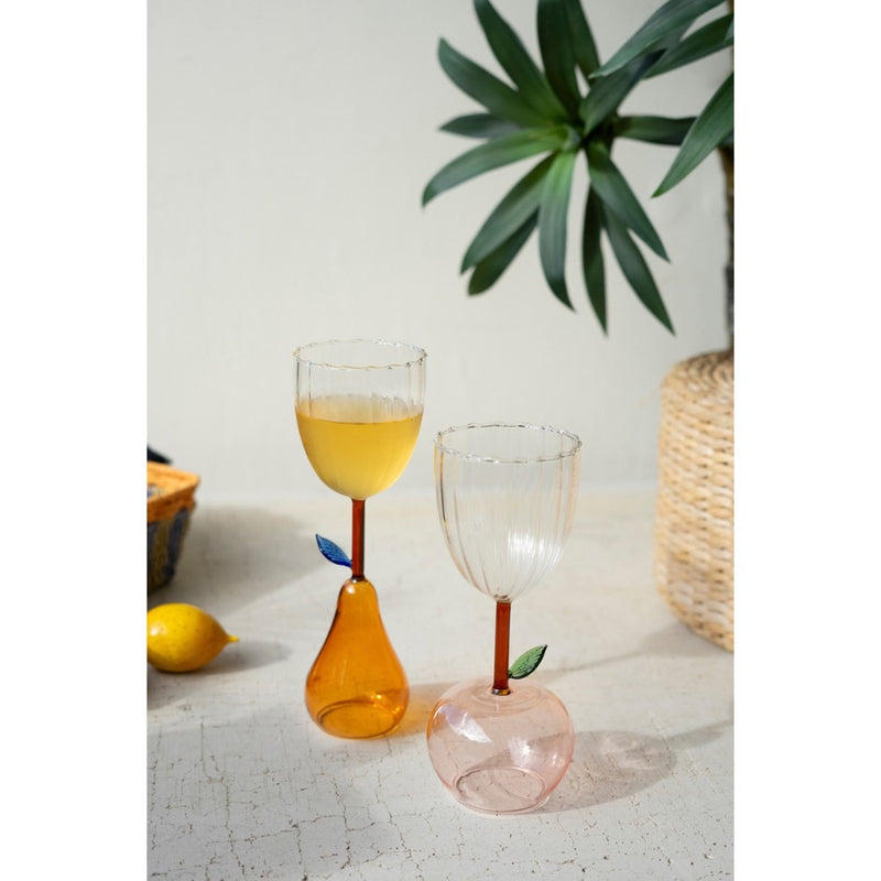 Two Kalalou Apple Base Wine Glasses with colored stems sit on a white table near a lemon and potted plant.