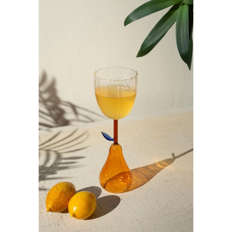 Kalalou's Pear Base Wine Glass with yellow liquid, leaf detail, two lemons, and plant shadows.