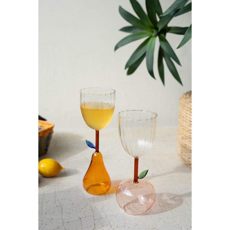 Two Kalalou pear base wine glasses, one with yellow liquid, sit on a table with a lemon and a plant.