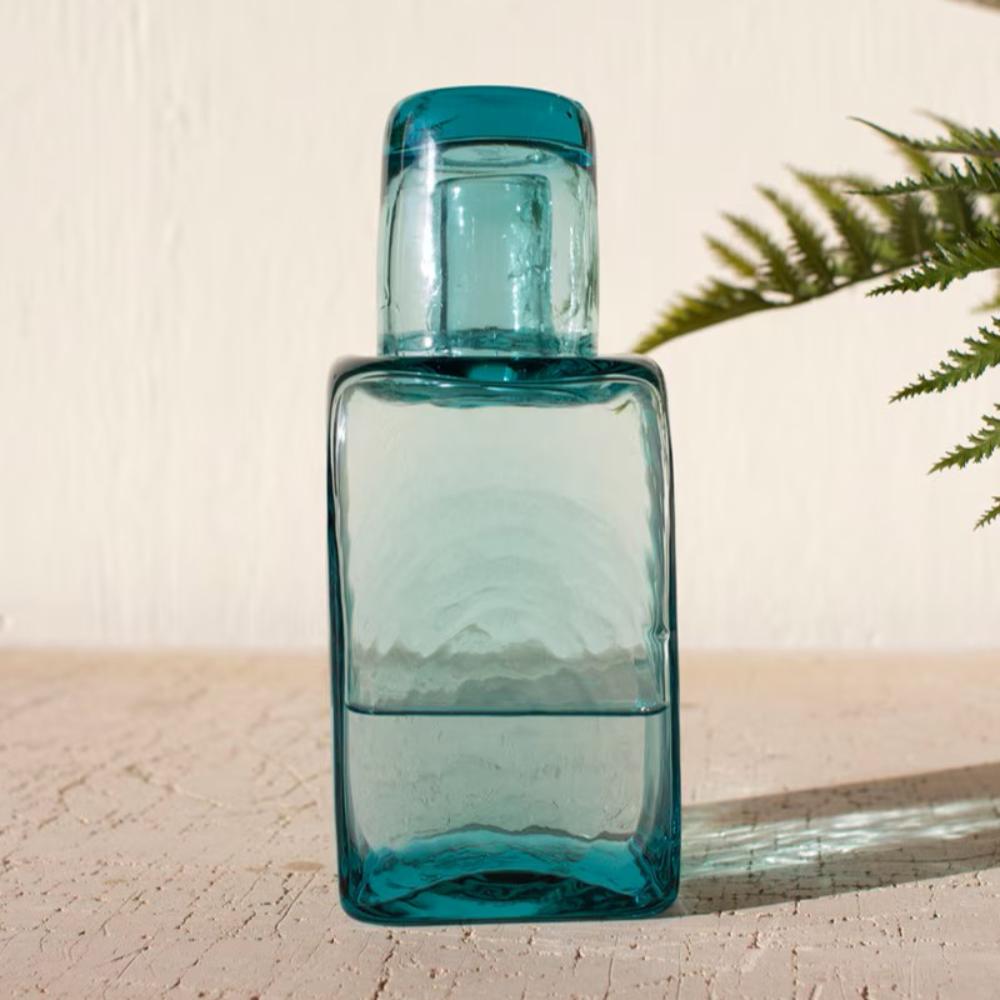 A translucent teal blue glass bottle with a matching screw-on cap, standing on a light textured surface against a cream wall with a green fern leaf in the background.
