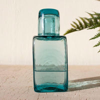 A translucent teal blue glass bottle with a matching screw-on cap, standing on a light textured surface against a cream wall with a green fern leaf in the background.